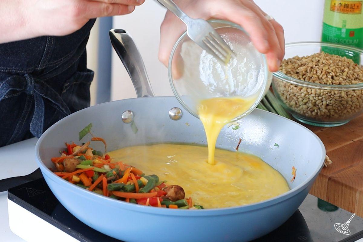 Someone pouring liquid eggs into a large pan containing vegetables.