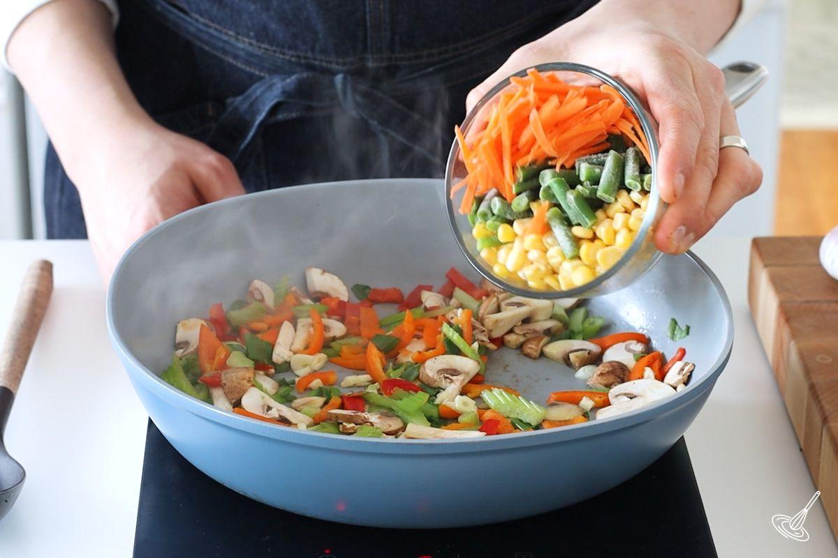 Someone adding vegetables to a large skillet of already cooking vegetables. 