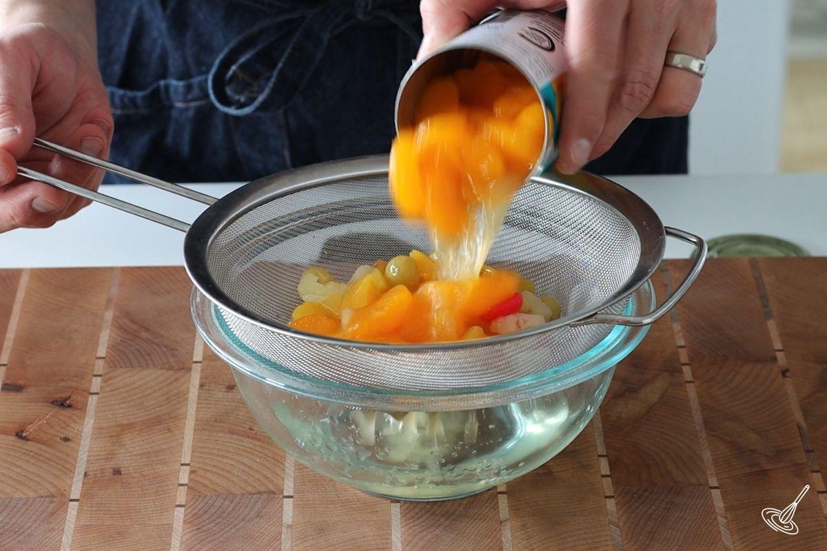 Someone adding cans of fruit cocktail to a fine mesh strainer. 