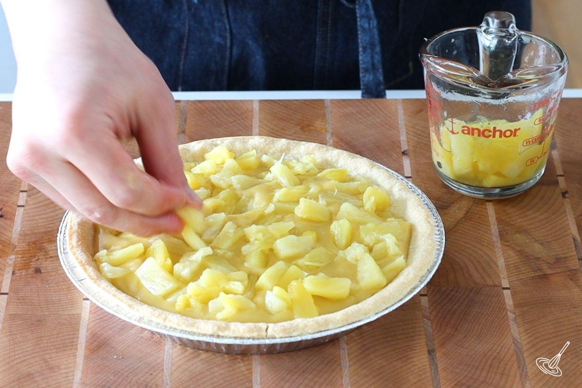 Someone placing pineapple pieces on top of a pie of banana pudding. 