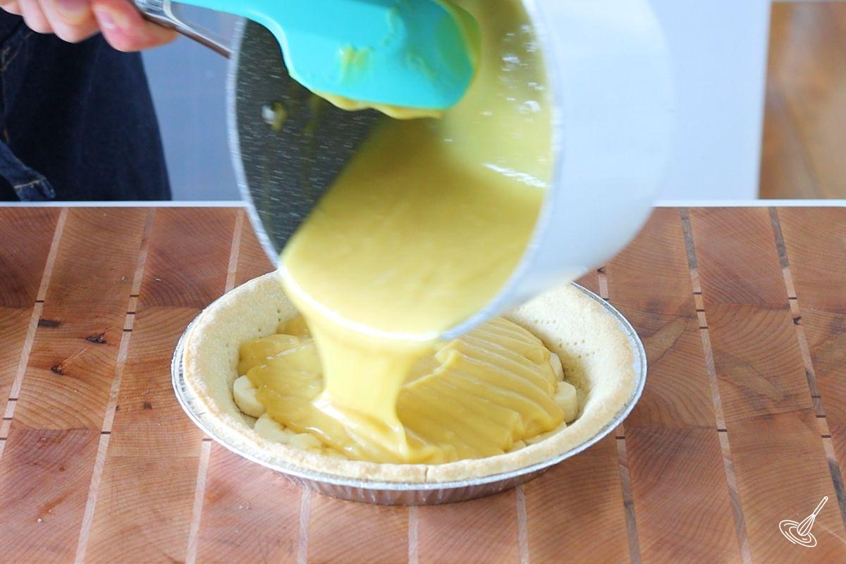 Someone pouring banana pudding oven a banana lined pie shell. 