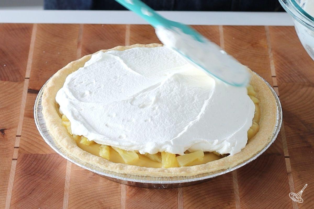 Someone using a rubber spatula to spread whipped cream over a banana split pie. 