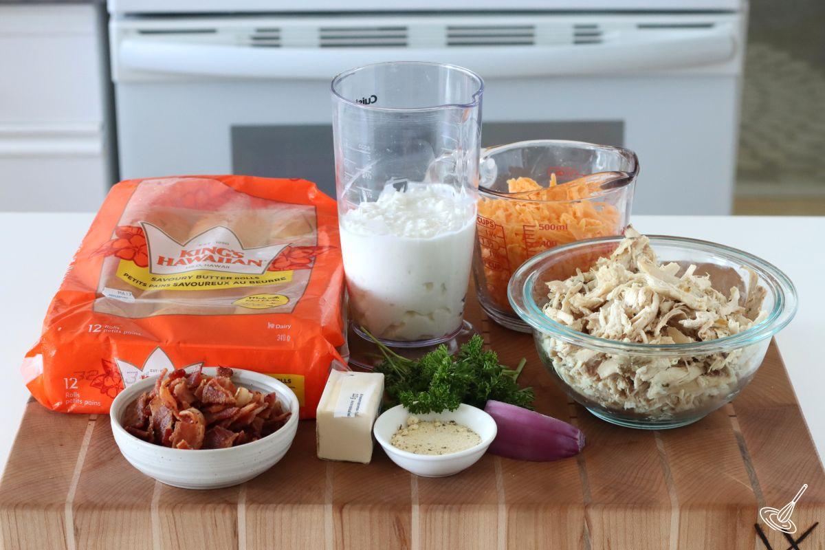 Ingredients on a cutting board including shredded chicken, grated cheese, cottage cheese, and Hawaiian rolls.