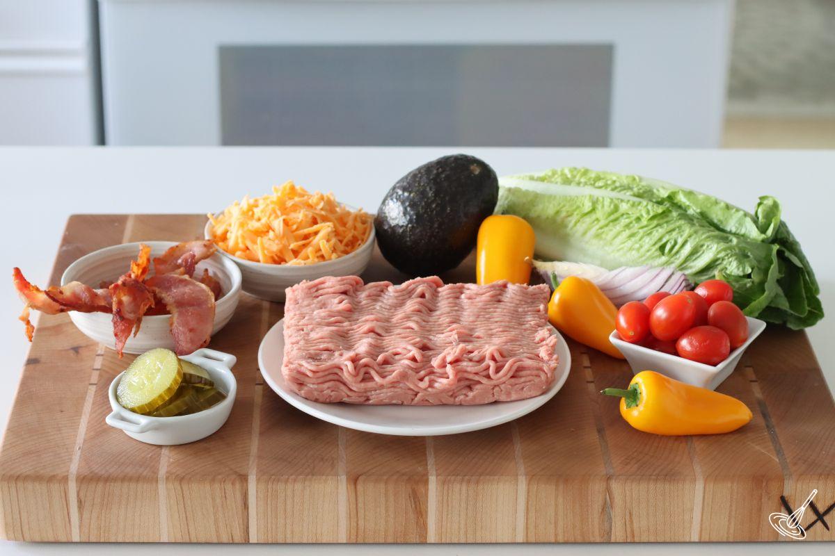 Ingredients on a cutting board, including romaine lettuce, tomatoes, bacon, pickles, and ground turkey.