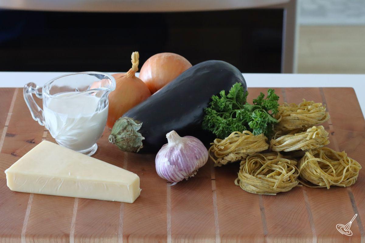 Ingredients on a cutting board including, aubergine, cheese, cream, garlic, and onion.