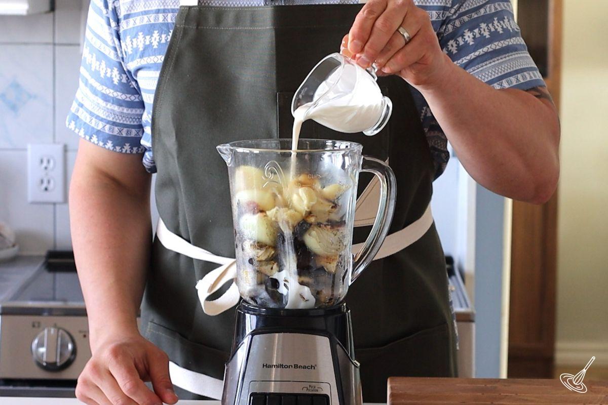 Someone adding cream to a blender with roasted vegetables.
