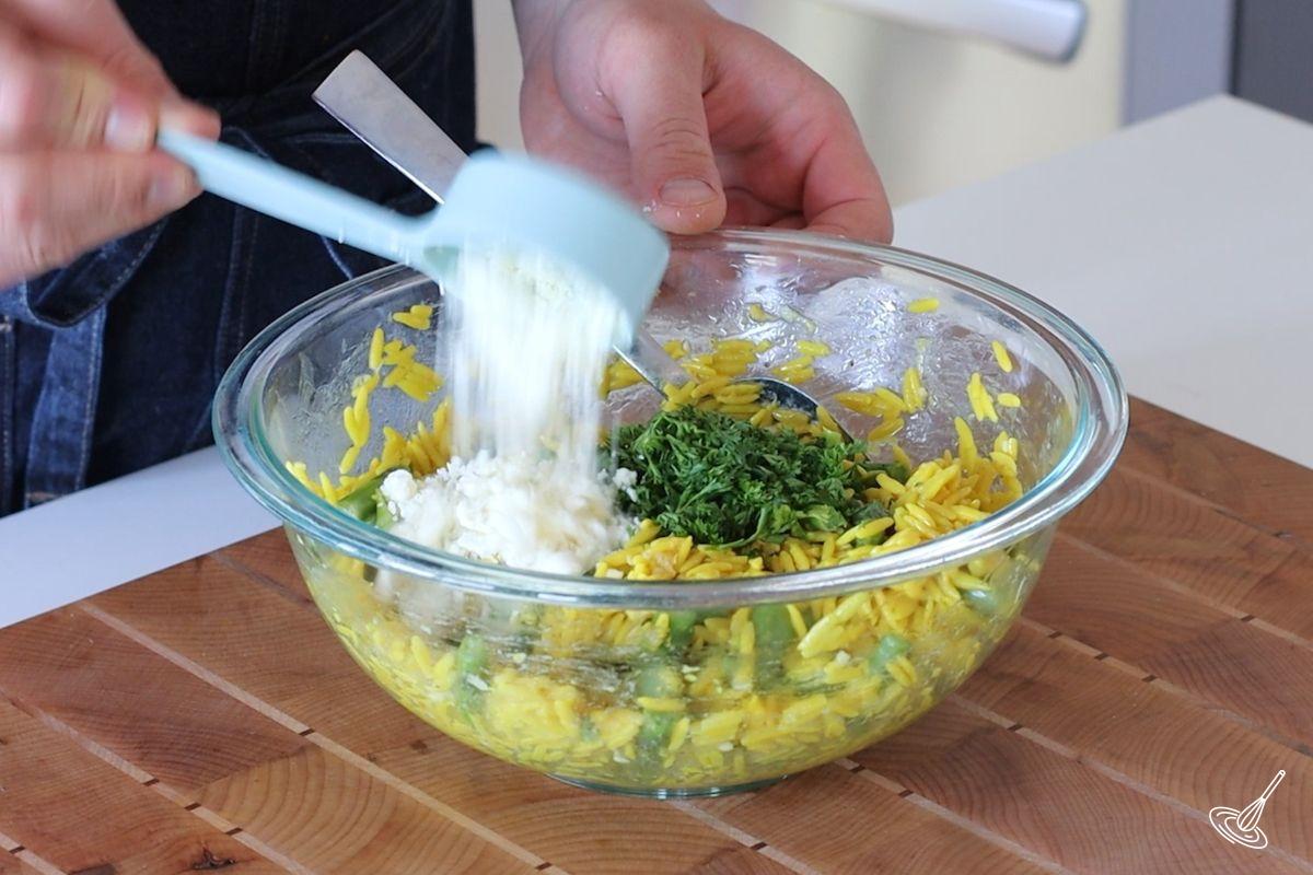 Someone adding crumbled feta cheese to a bowl of Lemon Asparagus Orzo salad.