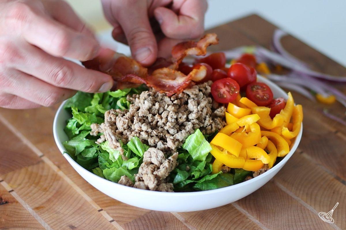 Someone adding slices of cooked bacon to a Turkey Burger Salad.