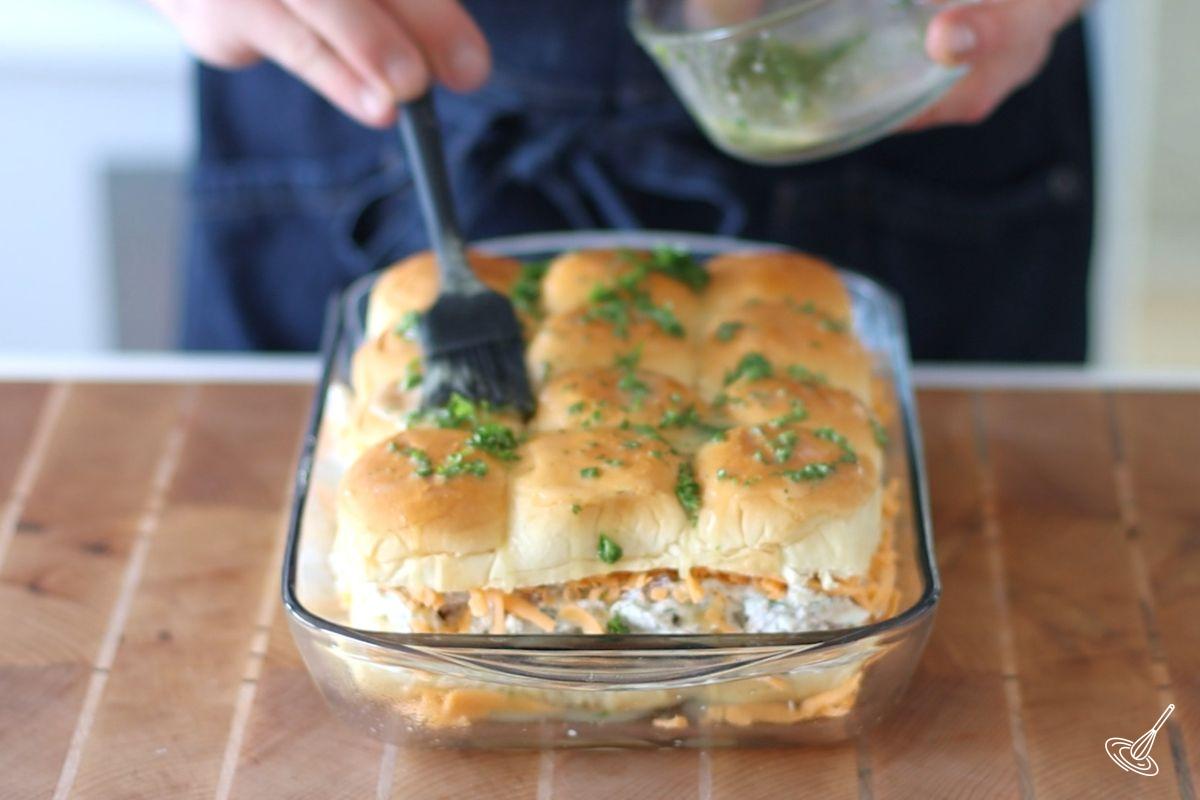 Someone brushing melted butter over the top of the crack chicken sliders.