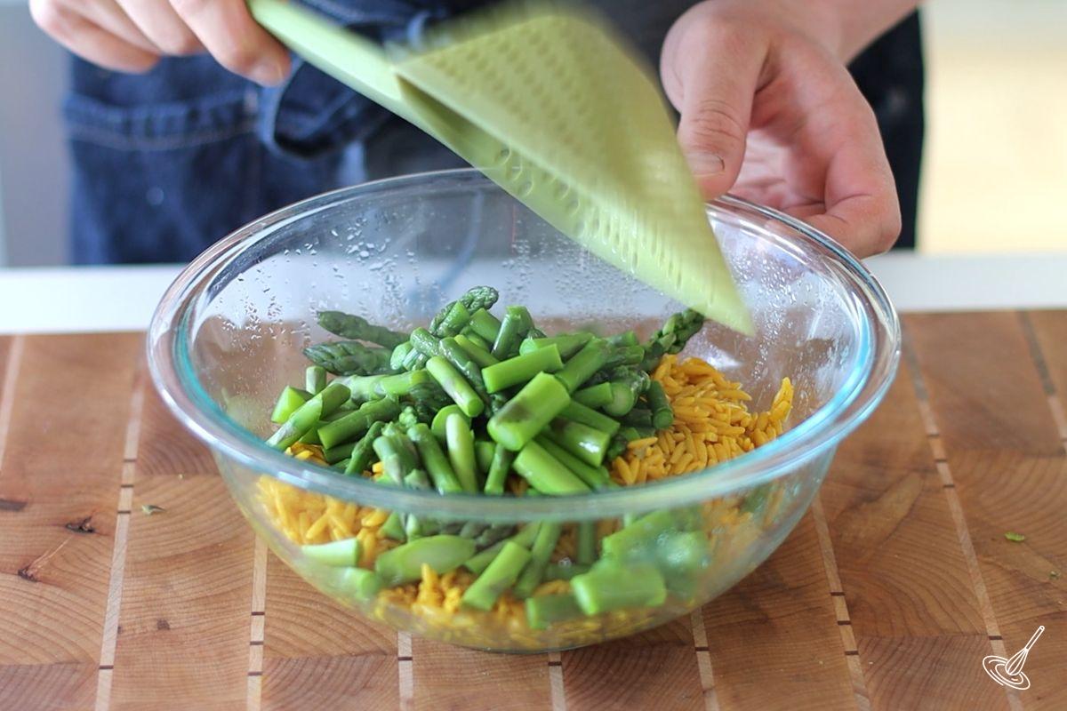 Someone adding blanched pieces of asparagus to a large bowl of cooked orzo.