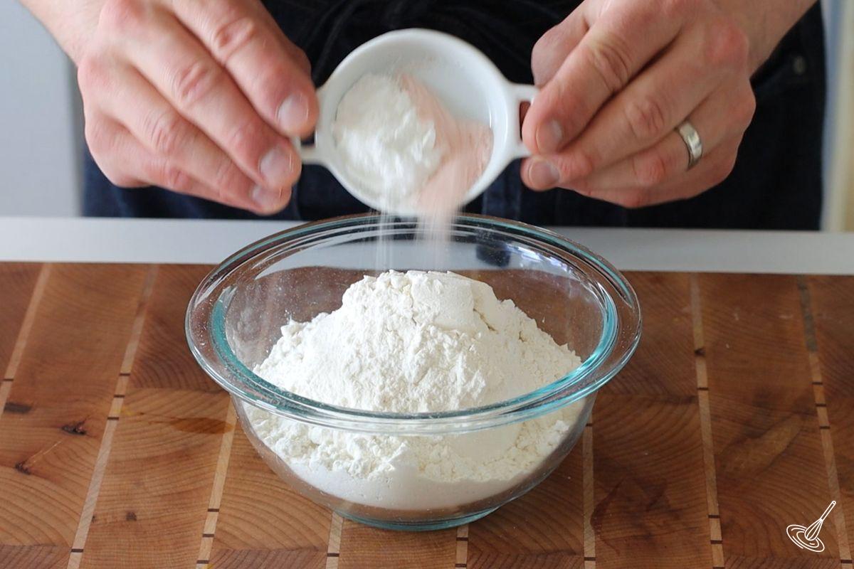 Someone adding baking powder to a small bowl of flour.