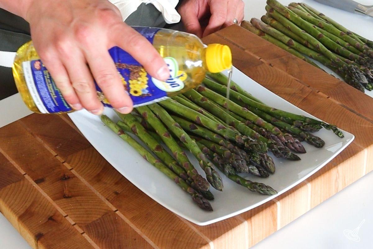 Someone drizzling cooking oil over a plate of asparagus. 