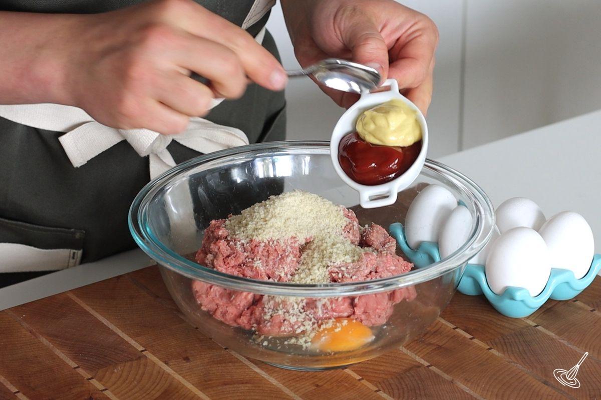 Someone adding mustard and ketchup to a bowl of meat mixture. 