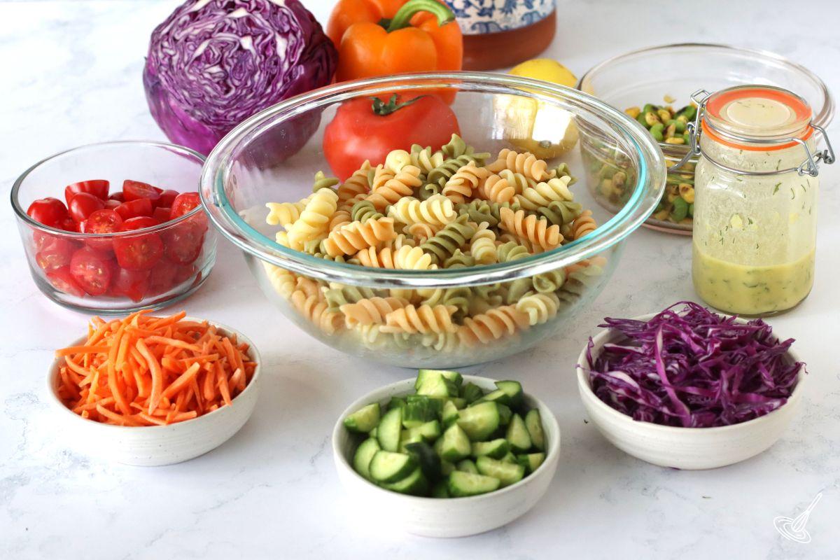 Ingredients on the table to make Rainbow Pasta Salad, including a bowl of cooked pasta, carrots, tomatoes, cucumbers, red cabbage. 