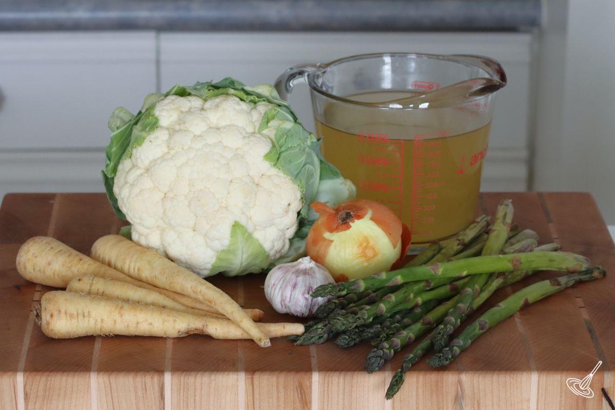 Ingredients on a cutting board including cauliflower, parsnips, asparagus, and chicken broth. 