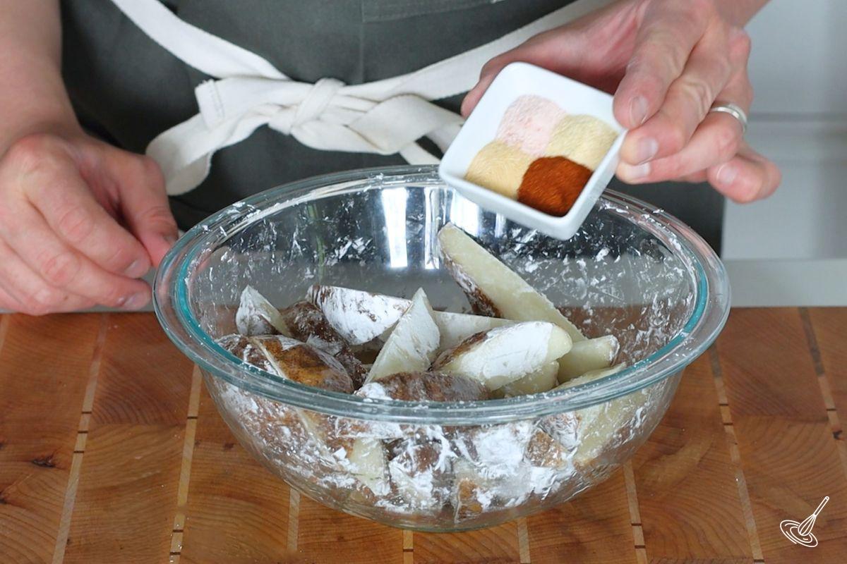 Someone adding spices to potato wedges in a bowl.