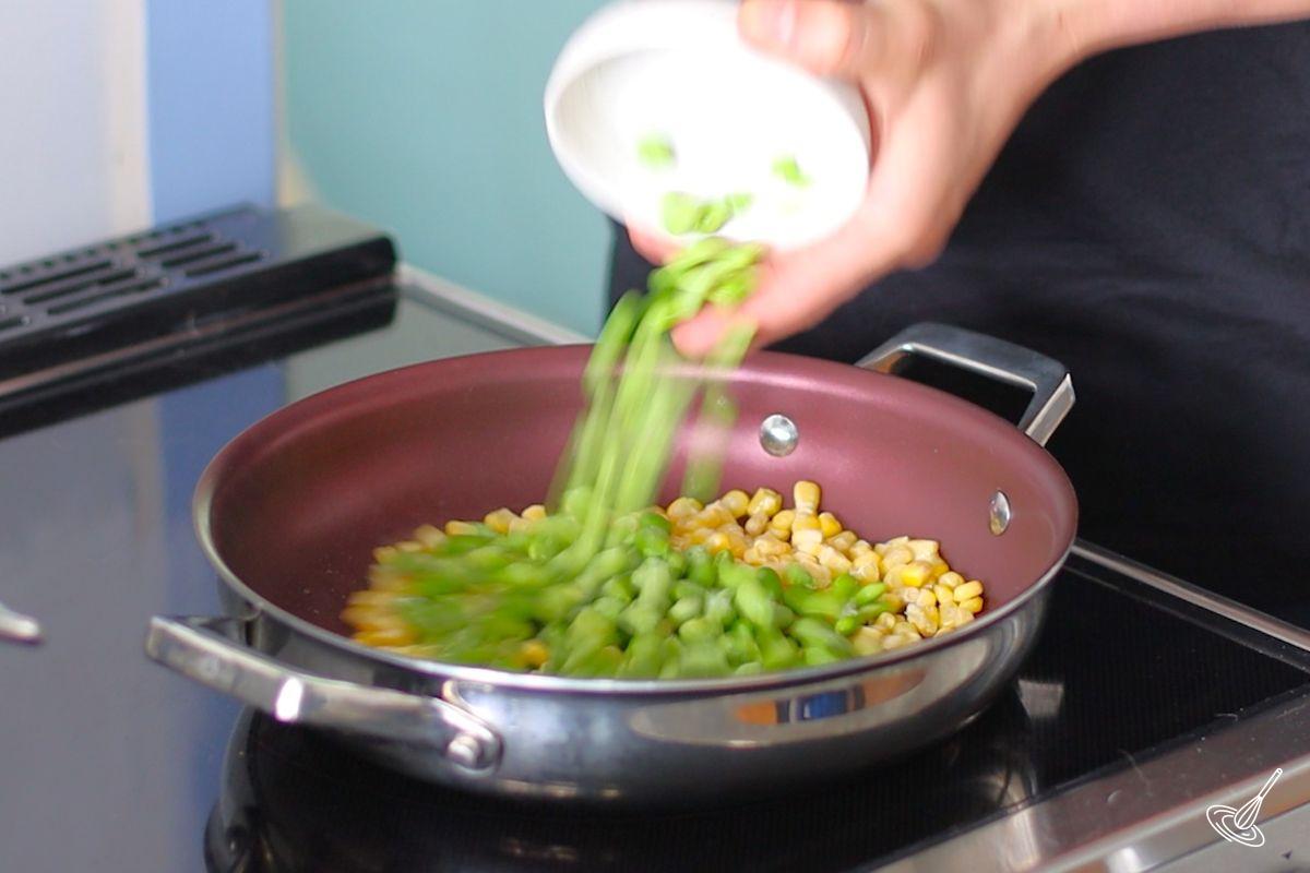 Someone adding edamame beans to a frying pan with corn kernels.