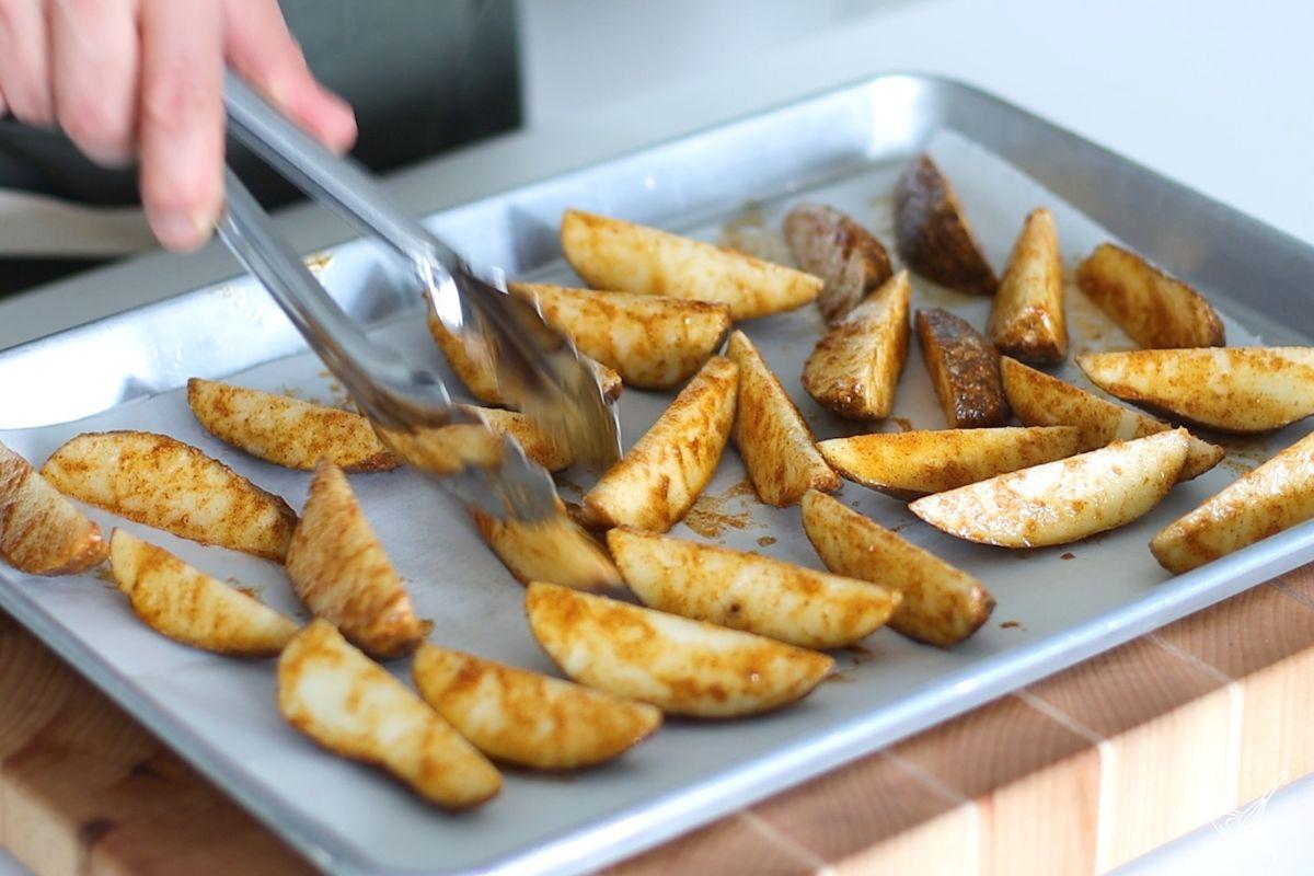 Someone using tongs to turn over potato wedges on a baking tray.