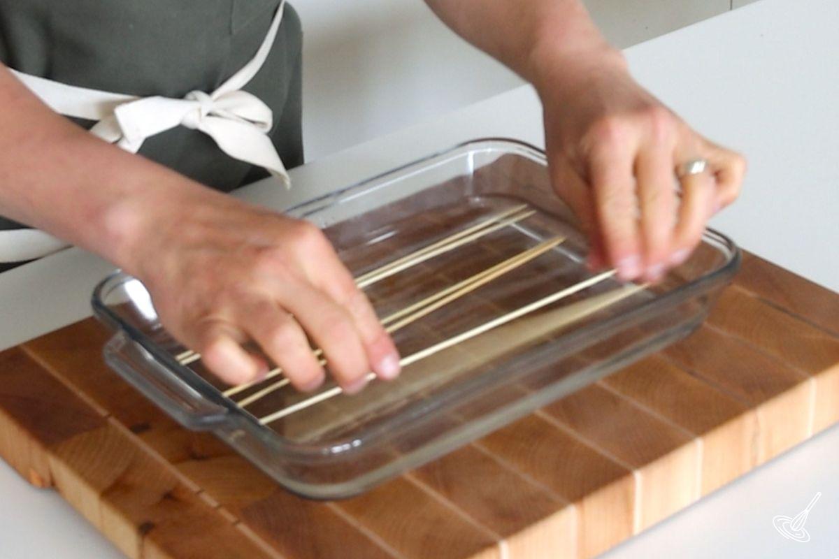 Someone placing wooden skewers sticks in a shallow dish of water.