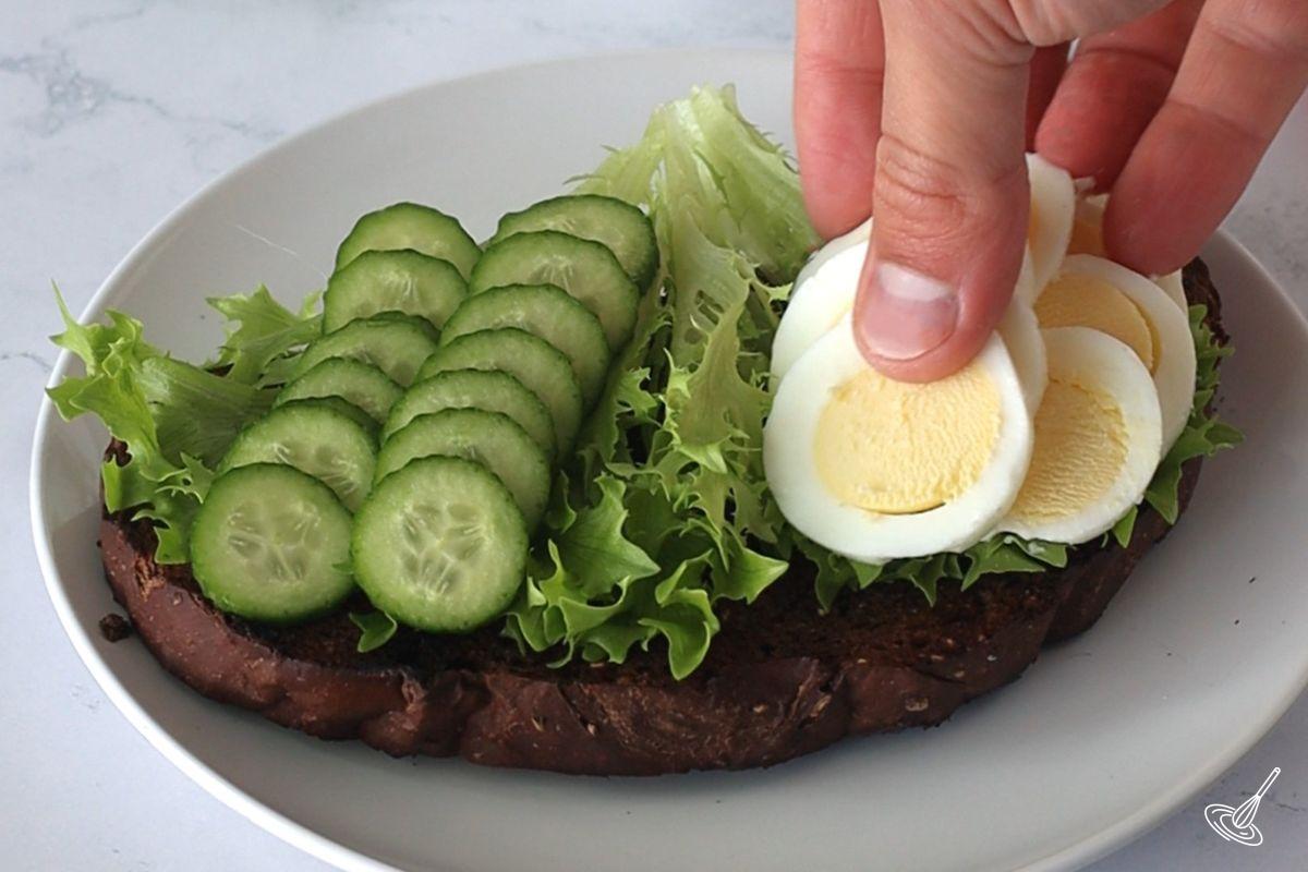 Someone placing sliced hard boiled eggs on a toast with lettuce and cucumber.