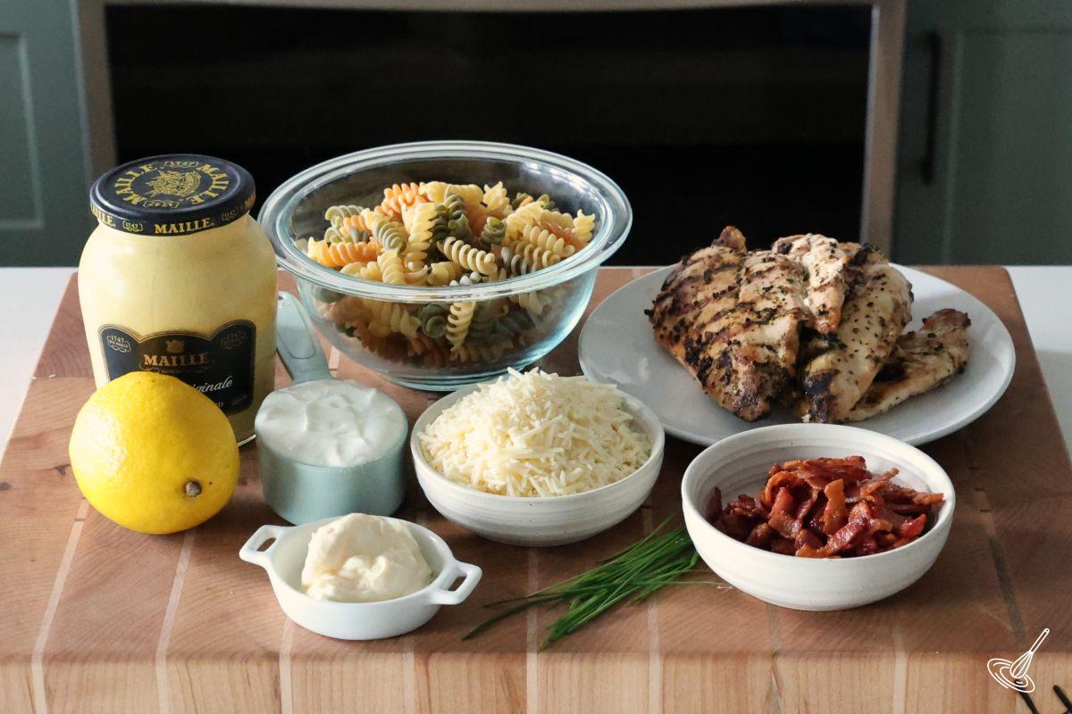 Ingredients on a cutting board, including chicken, bacon, pasta, parmesan cheese.