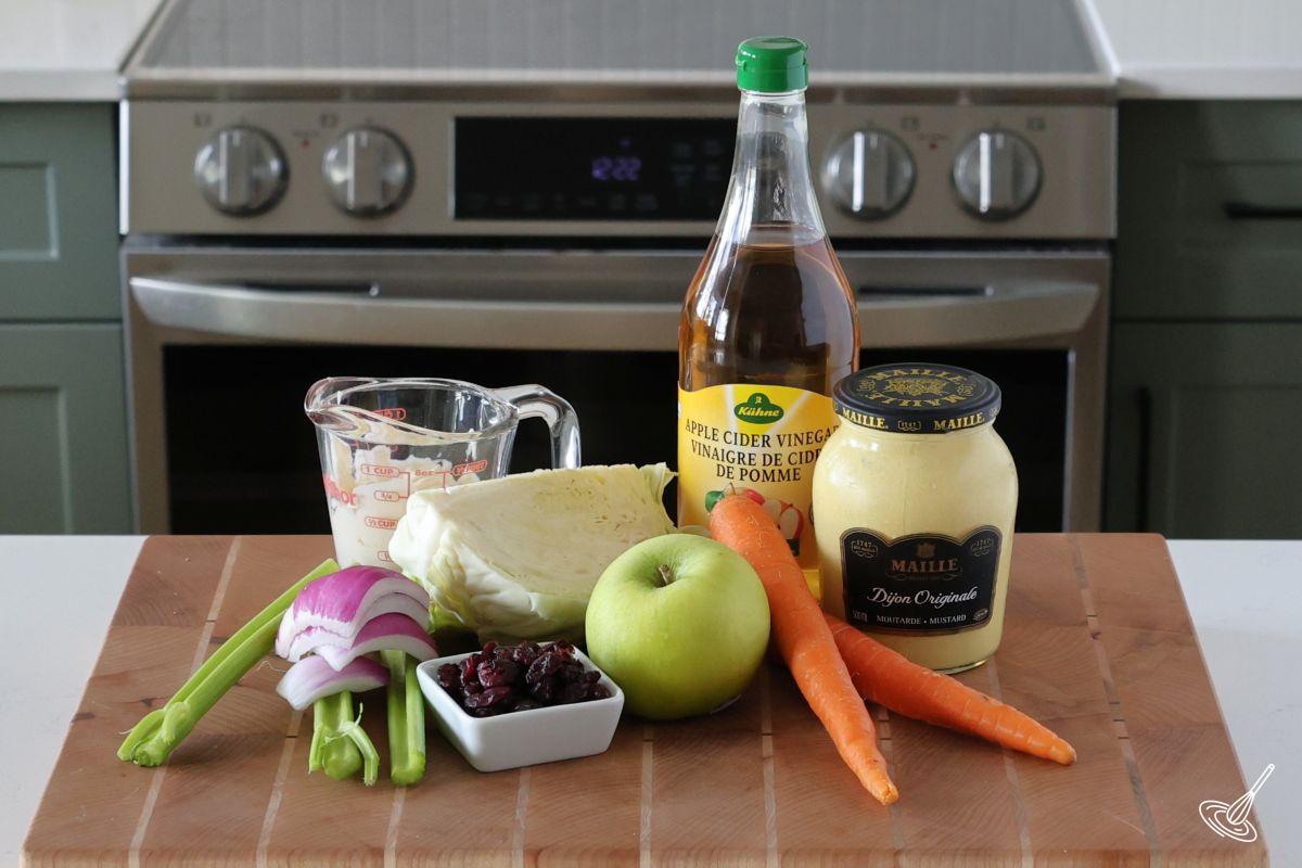 Ingredients on a cutting board including cabbage, apple, carrots, mayo, and cranberries. 
