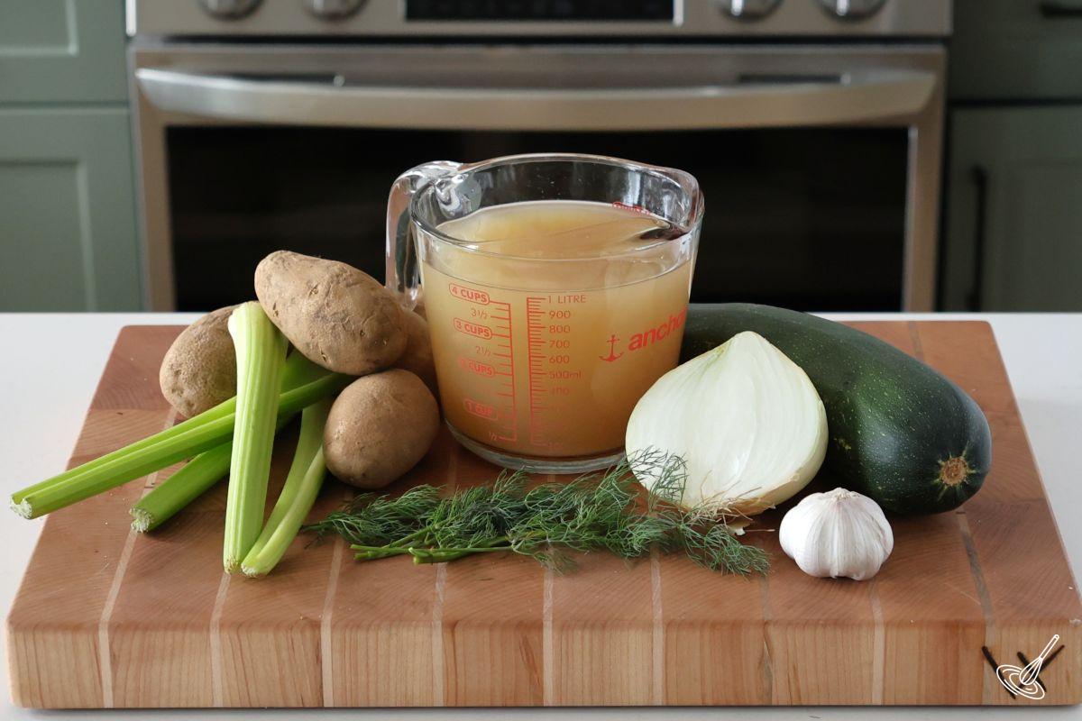 Ingredients on a cutting board, including chicken broth, zucchini onion, potatos and dill. 