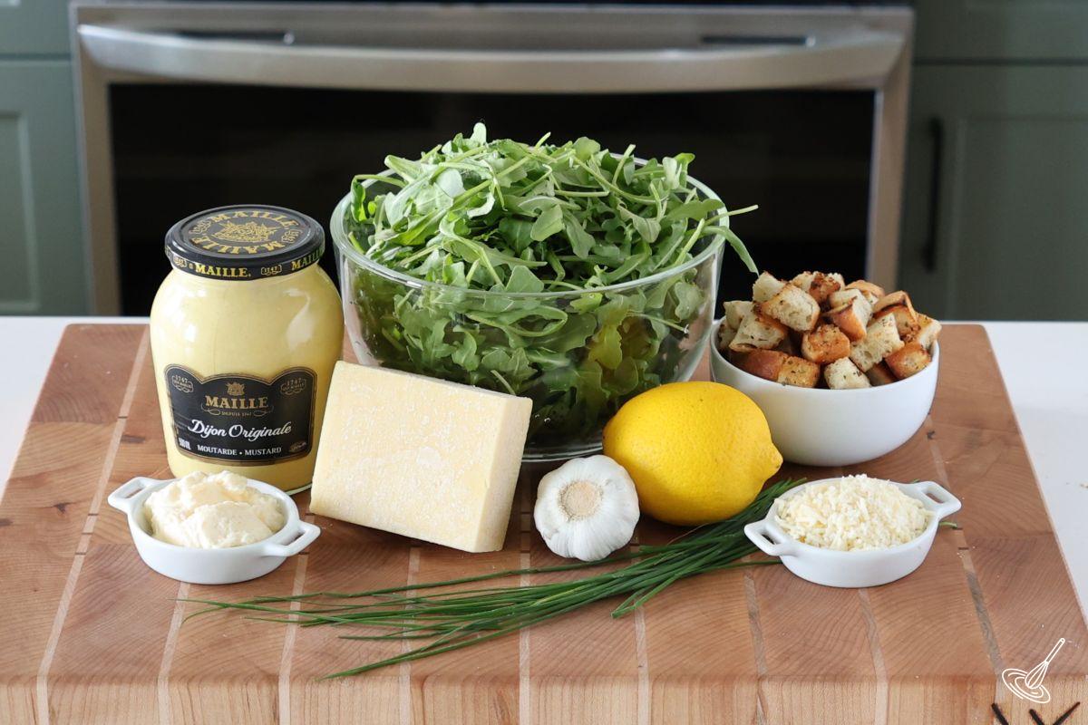 Ingredients on a cutting board, including baby arugula, croutons, lemon, parmesan cheese, garlic, and Dijon mustard. 