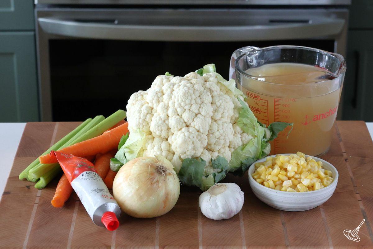 Ingredients on a cutting board, including chicken broth, cauliflower head, garlic, onion, and celery.