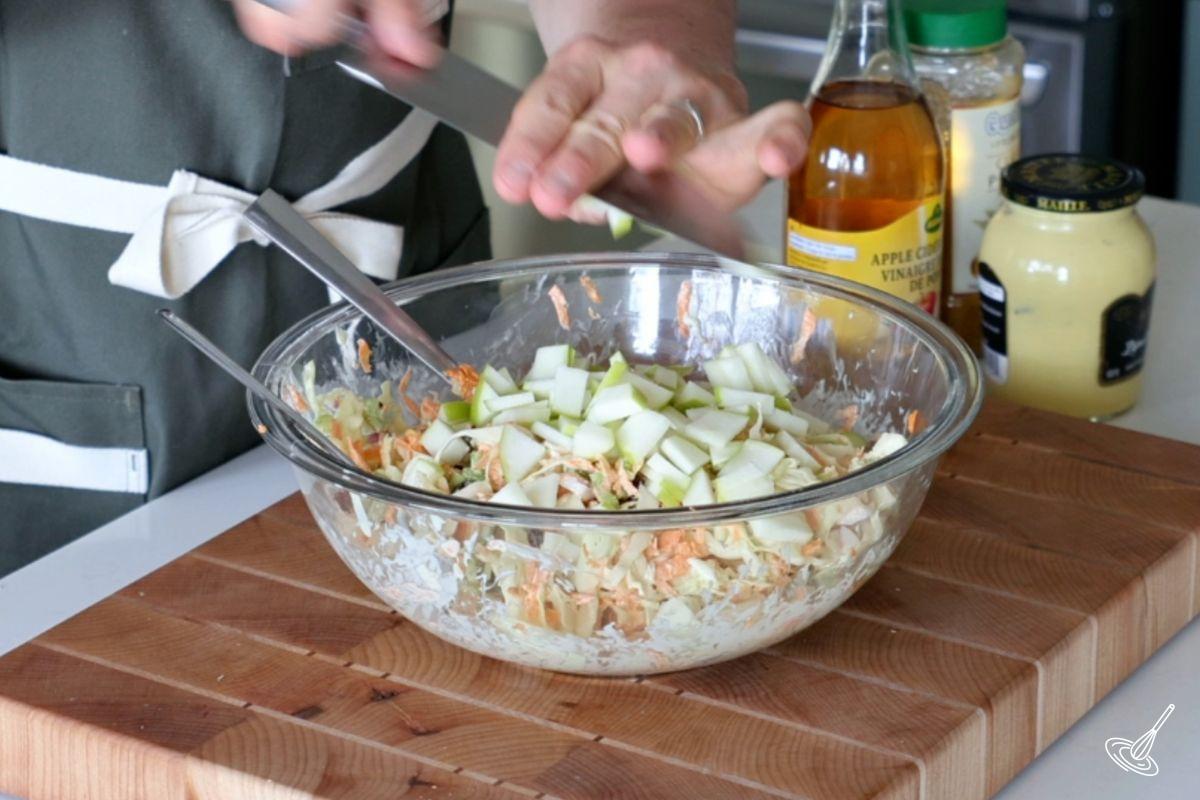 Someone adding chopped apples to a large bowl of coleslaw. 