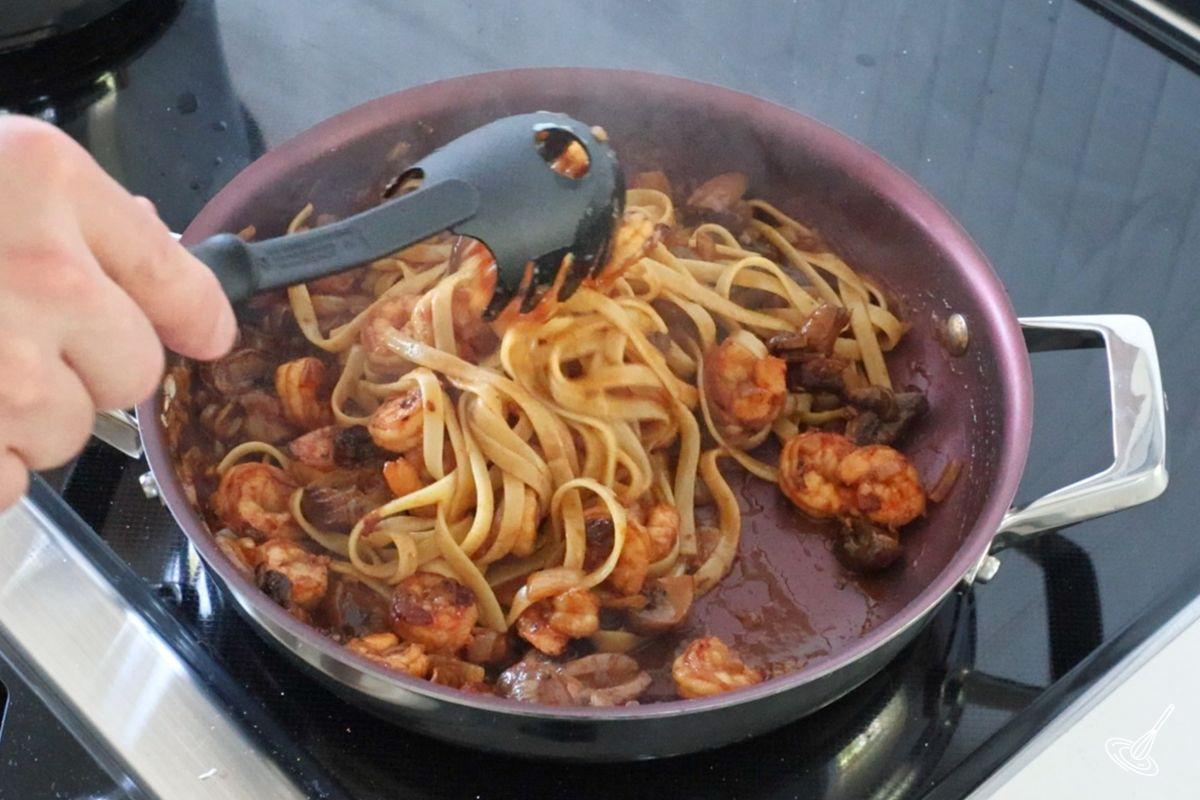 Someone stirring cooking pasta into a pan of shrimp and mushroom.