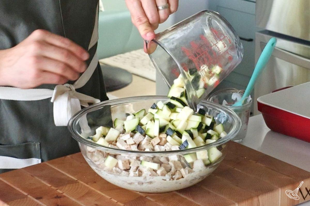 Someone adding cubed zucchini to a large bowl of chicken and cream mixture.