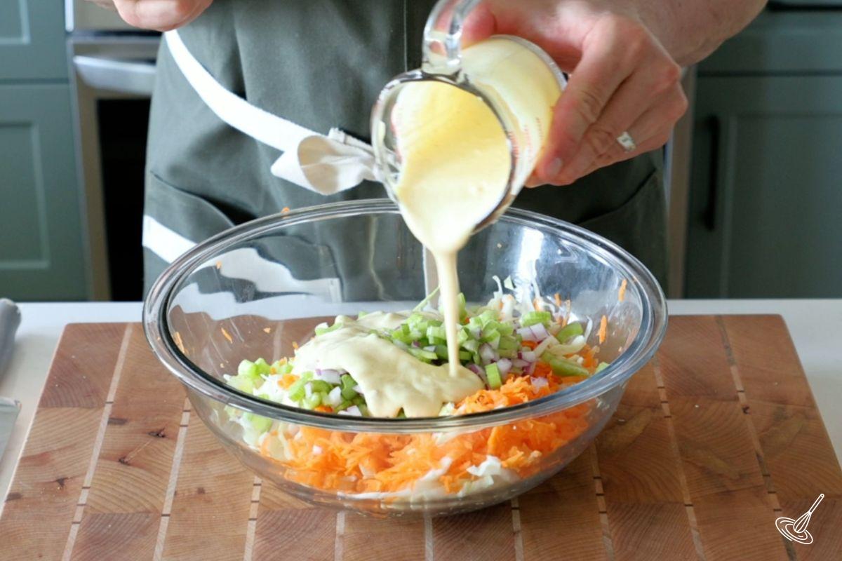 Someone pouring coleslaw dressing on top of Apple Cranberry Coleslaw.