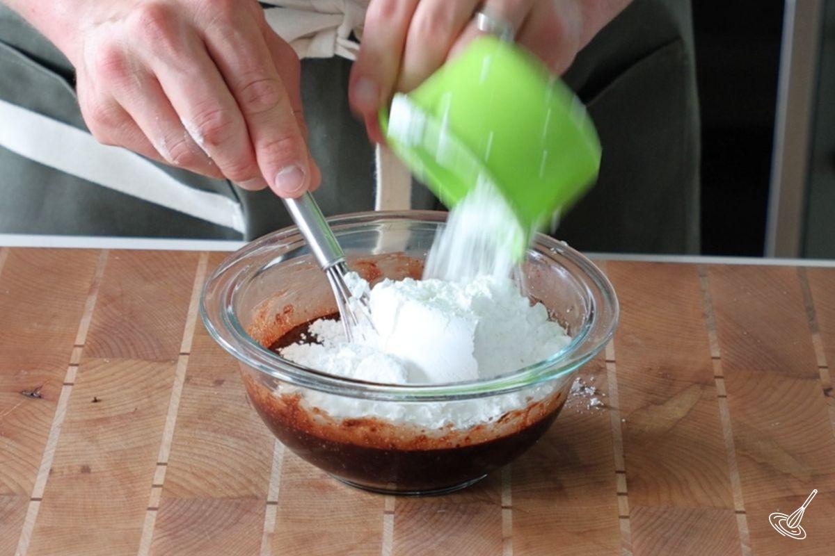 Someone adding a cup of icing sugar to a small bowl of chocolate icing. 