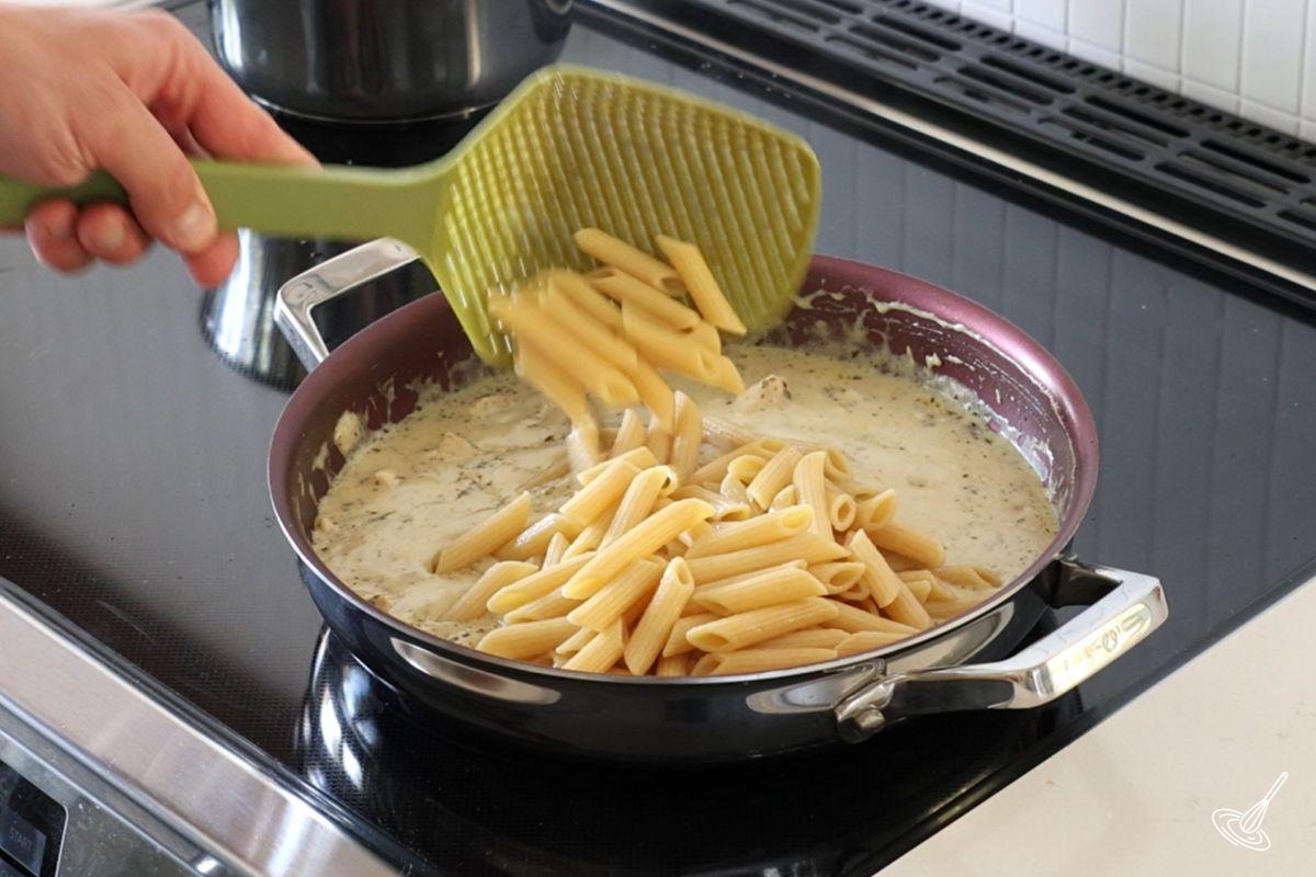 Someone adding cooked pasta to a large skillet of chicken alfredo sauce.