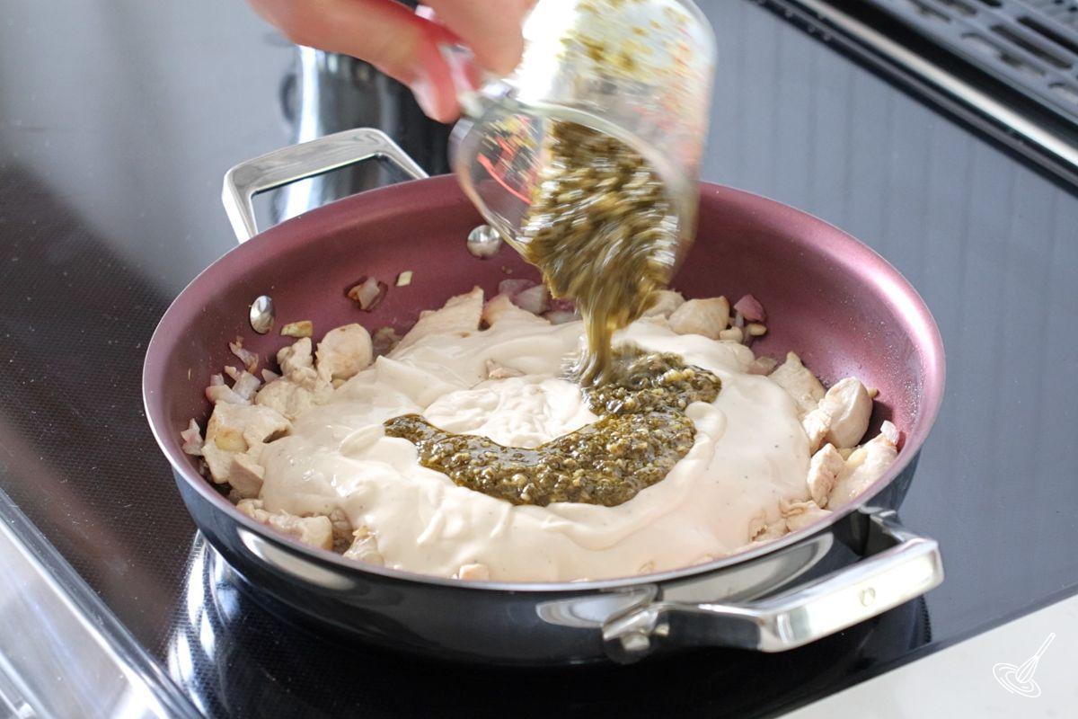 Someone adding pesto to alfredo sauce in a large pan. 