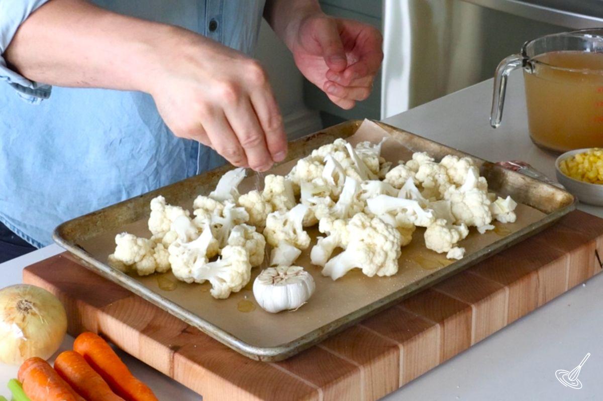 Someone sprinkling salt over a baking sheet of cauliflower florets.