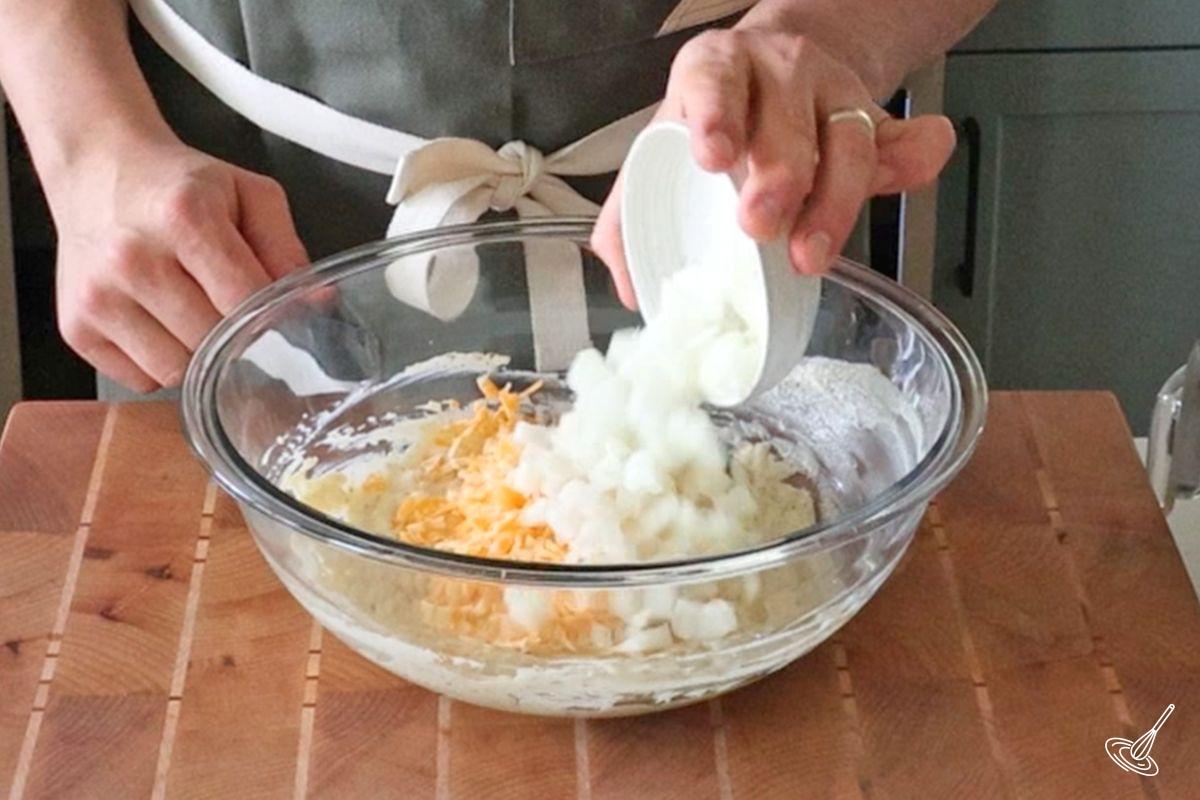 Someone added chopped onion to a large bowl of grated cheese and Greek yogurt. 