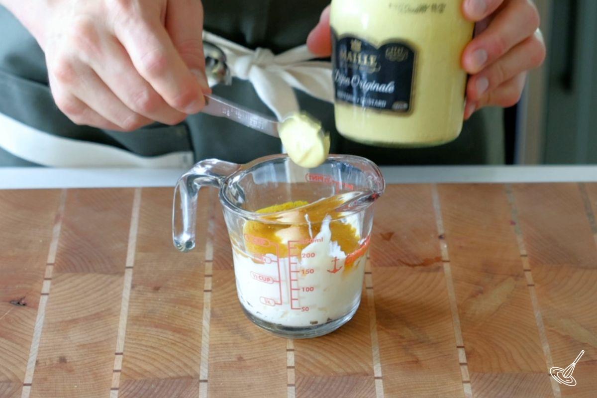 Someone adding Dijon mustard to a measuring cup of coleslaw dressing.