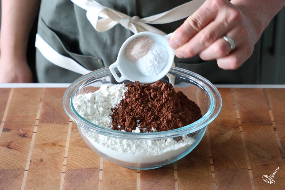 Someone adding baking powder to a small bowl of flour and cocoa powder.