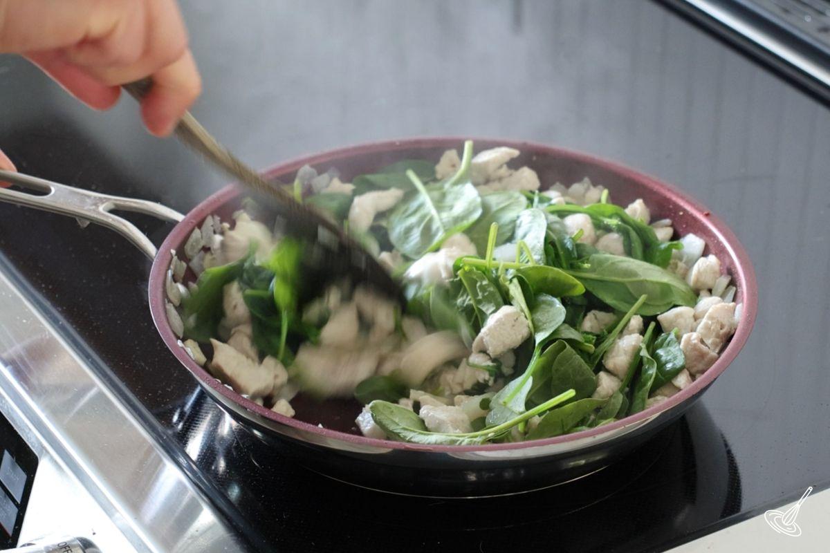 Someone stirring spinach into a large skillet with cooked chicken. 