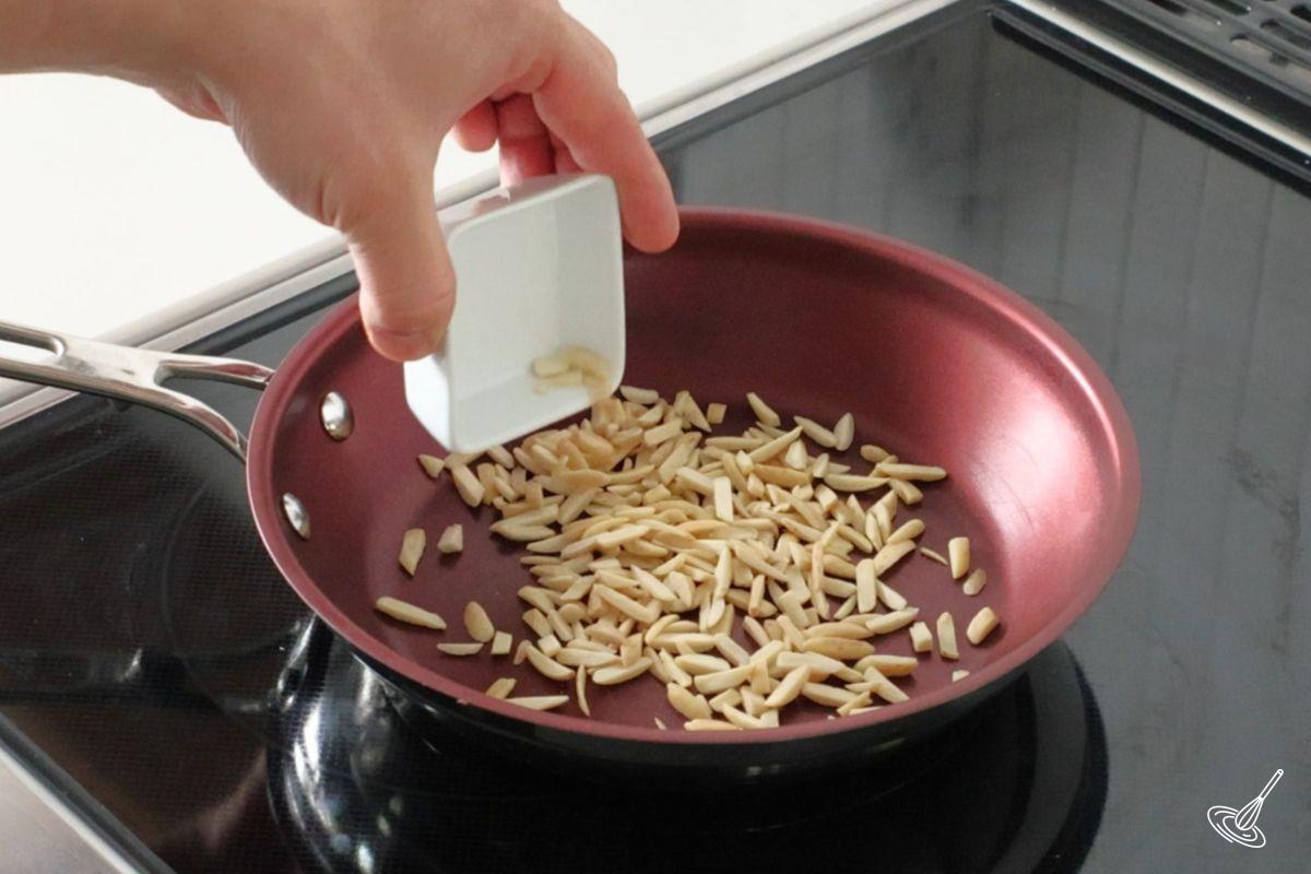 Someone adding slivered almonds to a frying pan. 