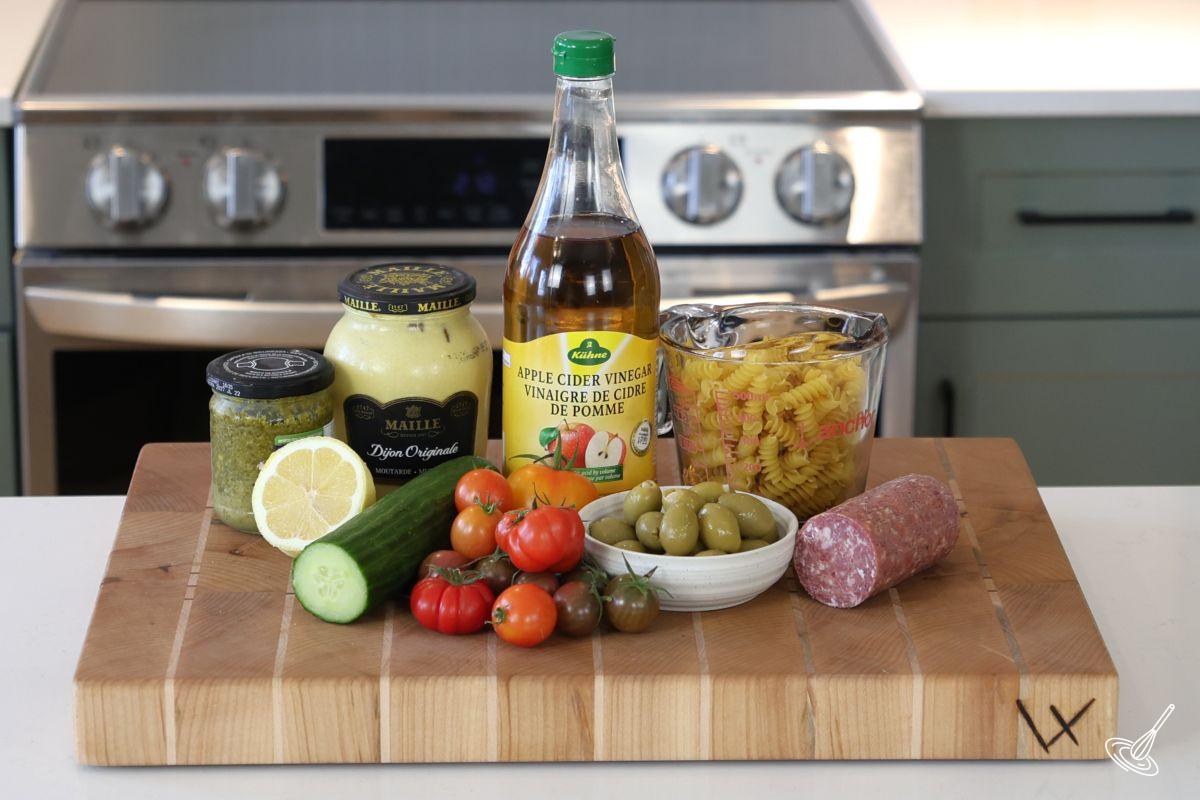 Ingredients on a wooden cutting board including pasta salami, cucumbers and tomatoes.