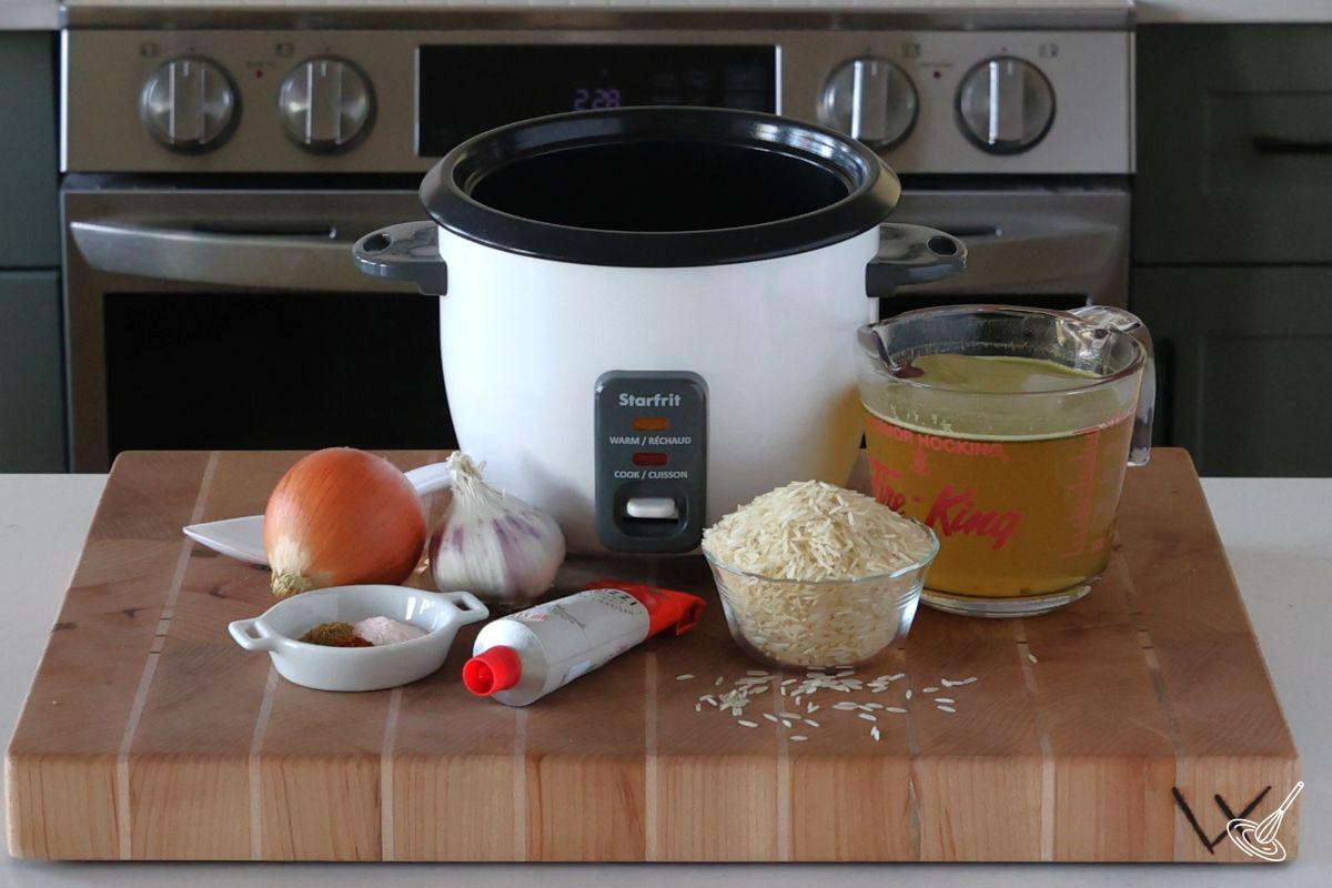 A rice cooker on a wooden cutting board, surrounded by ingredients to make the mexican rice.