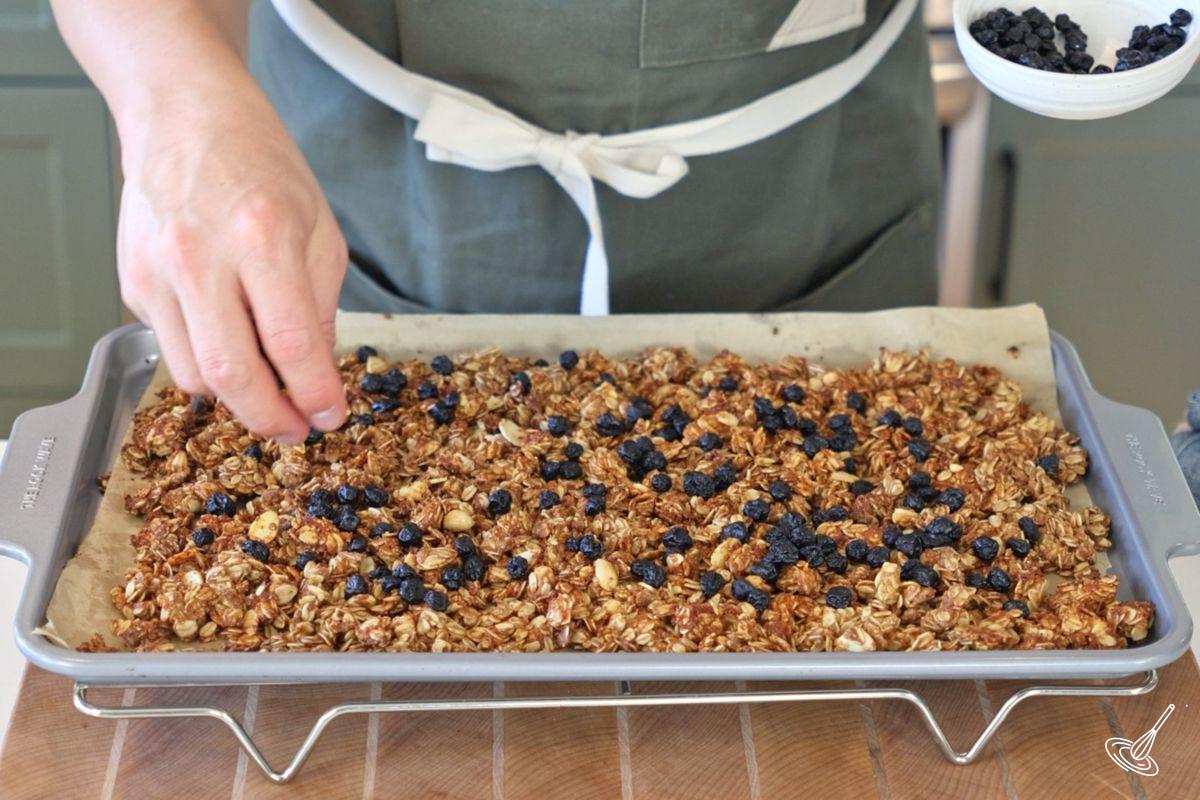 Someone sprinkling dried blueberries over a baked granola.