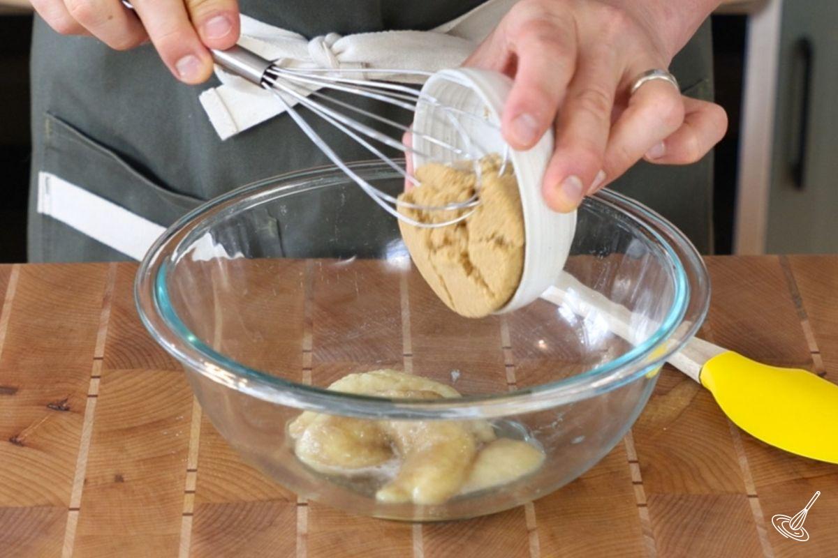 Someone adding brown sugar to a large glass bowl with banana puree. 