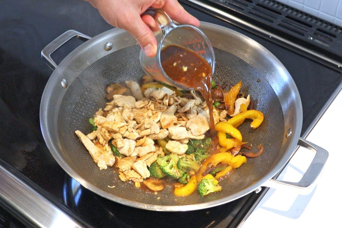 Someone pouring stir fry sauce into a work of cooked vegetables and chicken. 