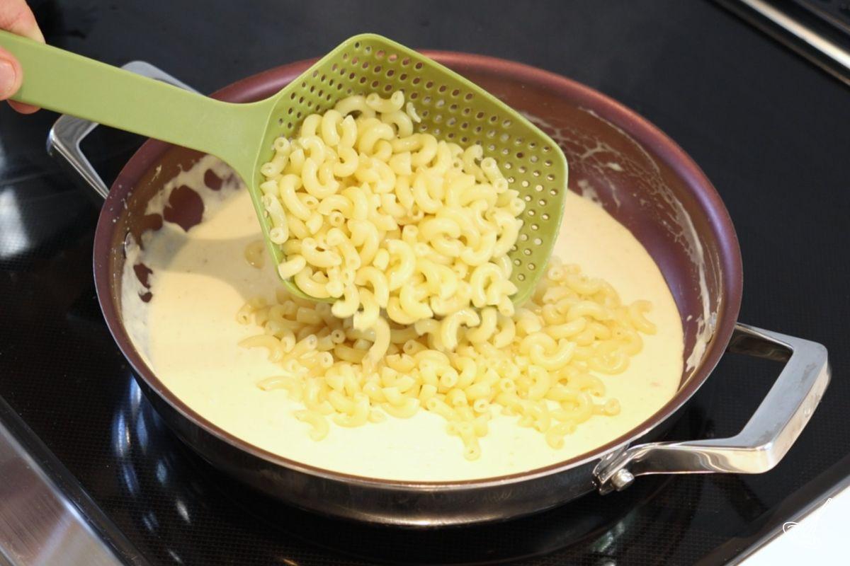 Someone adding cooked macaroni pasta to a sauce pan of cheese sauce.
