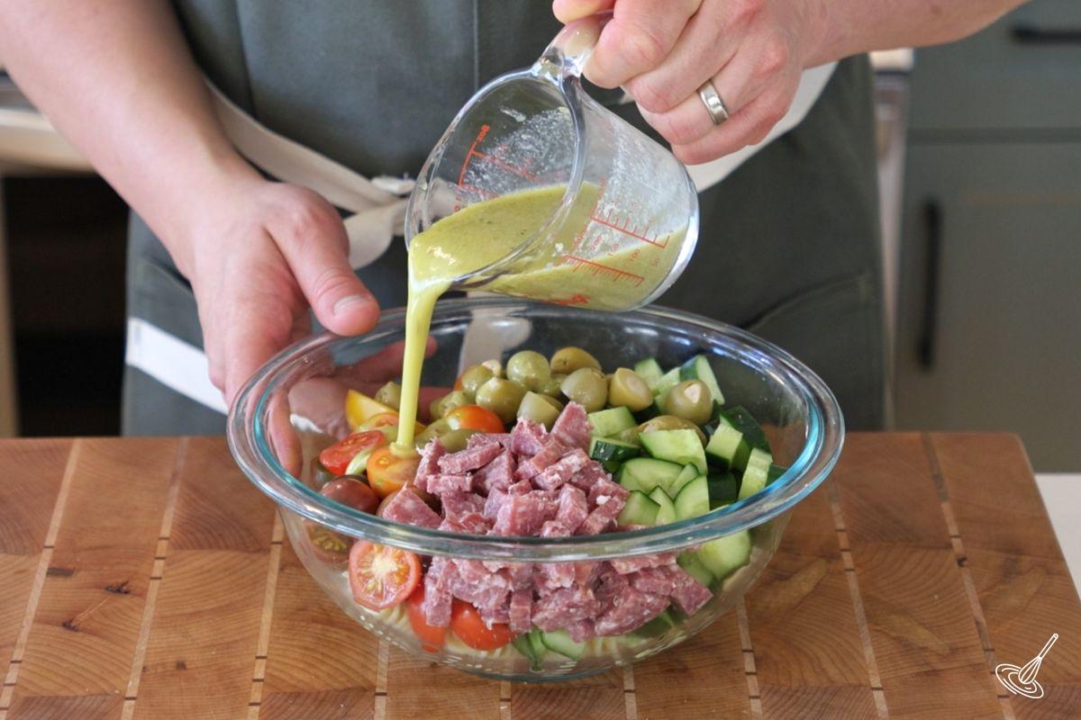Someone pouring salad dressing over a bowl of cucumber tomato pasta salad.
