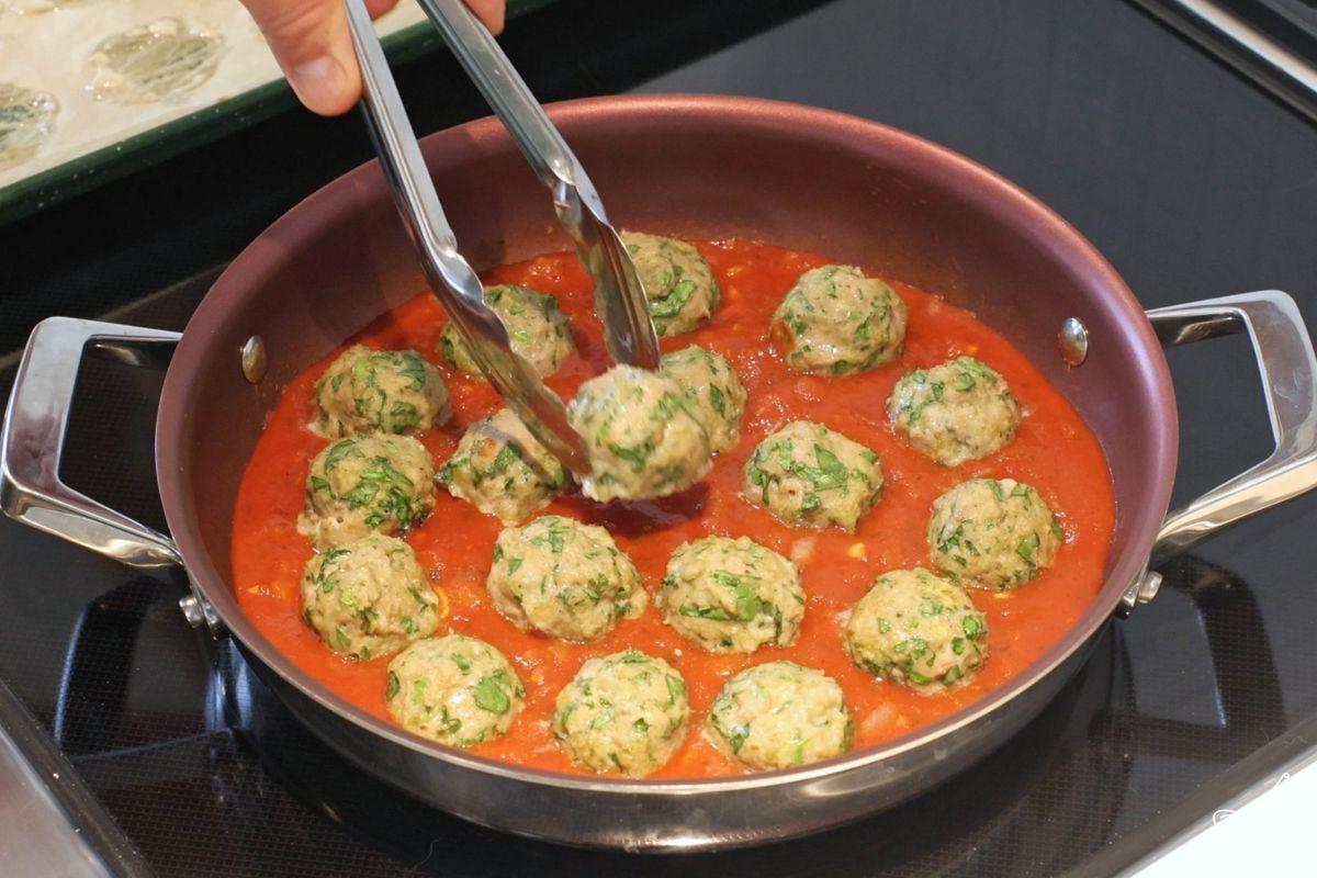 Someone adding meatballs to a large skillet containing tomato sauce.